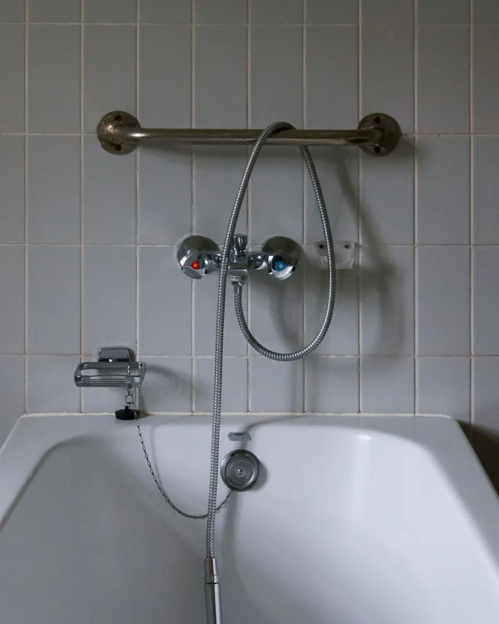 Older white tub against gray square tile with grab bar and hand shower